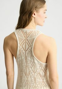 Top senza maniche in beige con un intricato motivo in pizzo, caratterizzato da un design con schiena bassa e un aspetto testurizzato e traspirante.