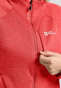 Rote Jacke mit strukturiertem Oberflächenmaterial, Frontreißverschluss und einer seitlichen Reißverschlusstasche. Mit einem Logo im Brustbereich.