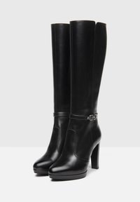 Bottes en cuir noir, arrivant au genou, avec une finition lisse, bout arrondi et un talon haut ; dotées d'une sangle argentée autour de la cheville.