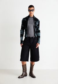 Person wearing black and gray tie-dye zip-up sweater, gray fitted top, black knee-length wide shorts, brown patterned cowboy boots, and dark sunglasses.