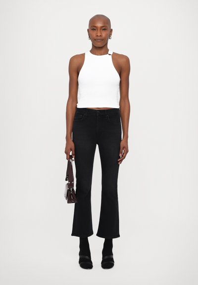 Top alb fără mâneci, blugi negri evazați, cizme negre până la glezne, ținând o geantă închisă la culoare. Material neted, design ajustat, estetic minimalist.