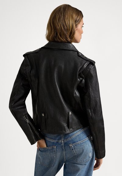 Crna kožna motoristička jakna s epauletima, uskim krojem i detaljima na patent zatvaraču; glatka tekstura i skraćena duljina. Nošena s plavim trapericama.