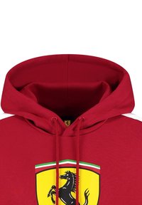 Czerwona bluza z kapturem z dużym czarnym logo Ferrari na żółtej tarczy, z białymi ukośnymi paskami na każdym ramieniu i sznurkiem do ściągania.