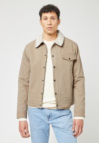 Giacca khaki con un colletto di peluche crema, bottoni a pressione e una tasca sul petto. Indossata sopra un maglione di lana crema e jeans azzurri chiari.