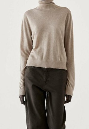 Pull à col roulé beige associé à un pantalon noir ample, présenté sur une personne devant un fond blanc uni.