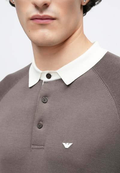 Polo marron avec un col blanc, trois boutons, et un petit logo sur la poitrine. Texture côtelée et détails contrastants aux épaules.