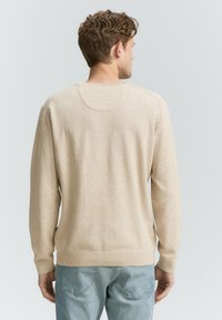 Mann von hinten gesehen, der einen beigen Strickpullover und helle blaue Jeans gegen einen schlichten hellen Hintergrund trägt.