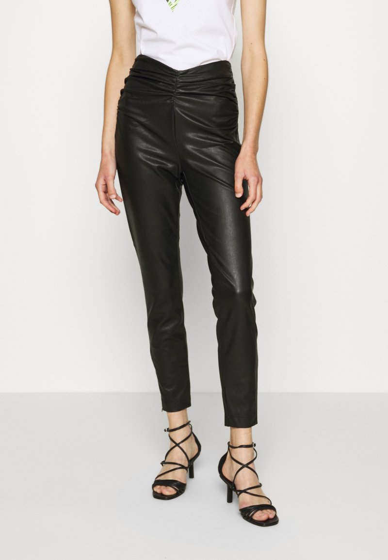 Černé kožené skinny kalhoty s řaseným páskem, doplněné černými sandály na vysokém podpatku s pásky. Hladká textura a těsně padnoucí design.