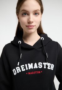 Fekete kapucnis pulóver zsinórral, fehér kiemelt betűzéssel a "DREIMASTER" felirattal és piros részletekkel. Puha anyag és laza dizájn.