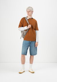 Brun t-shirt, lyse stribede langærmede skjorte indenunder, denimshorts, hvide sneakers med beige såler og en lys grå skuldertaske.
