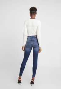 Tunna blå jeans med en slät textur, hög midja och detaljer på bakfickorna. Kombinerat med en vit långärmad topp.