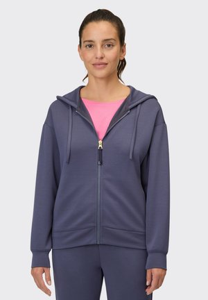 Woman with dark hair wears a purple zip-up hoodie over a bright pink shirt, standing against a plain light background.