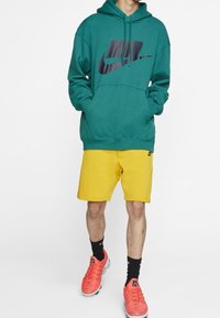 Teal huppari mustalla Nike-logolla, yhdistettynä keltaisiin shortseihin. Jalkineet ovat kirkkaan punaiset lenkkarit. Asusteessa on rento, urheilullinen muotoilu.
