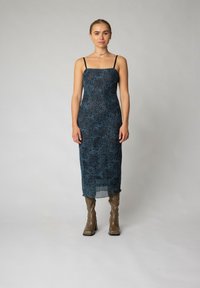 Rochie midi fără mâneci, de culoare albastru închis, cu o textură modelată; are bretele subțiri și o siluetă dreaptă, asociată cu cizme maro până la genunchi.