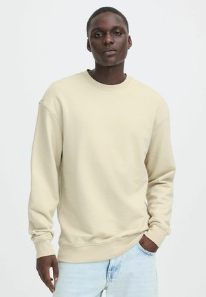 Giovane uomo con una felpa beige girocollo e jeans azzurri, in piedi con una mano in tasca su sfondo bianco semplice.