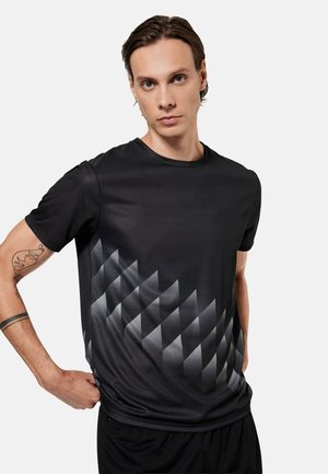 Ung mand med kort brunt hår iført en sort kortærmet skjorte med et gråt geometrisk diamantmønster foran, står mod en hvid baggrund.