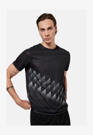 Jonge man met kort bruin haar draagt een zwart shirt met korte mouwen met een grijs geometrisch diamantpatroon aan de voorkant, staand tegen een witte achtergrond.