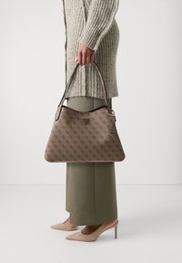 Brun mönstrad handväska med en triangulär logotyp, slät textur och ett axelband, hålls av en person som bär en ljus tröja och byxor.