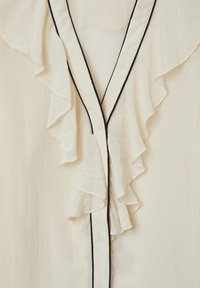 Blusa off-white con scollatura a V profonda, fronte arricciato con dettagli neri e chiusure a bottone. Il tessuto ha un aspetto testurizzato e stropicciato.