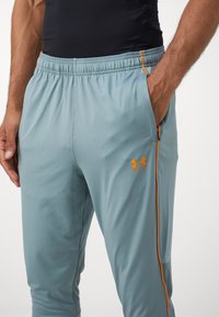 Svetlo modre športne hlače iz gladkega, elastičnega materiala. Imajo oranžno stransko črto in subtilen logotip Under Armour na nogi.