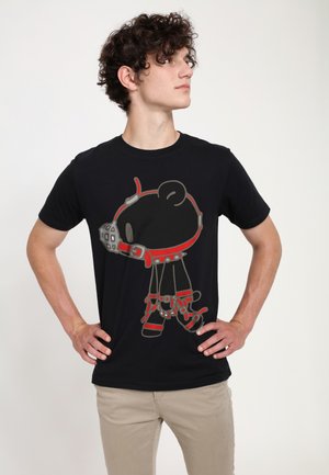 Ung mand med krøllet hår står med hænderne på hofterne iført en sort t-shirt med en tegneseriebjørn med rød sele og mundkurv.