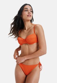 Set de bikini portocalie cu nervuri, ce prezintă un top balconet cu bretele ajustabile și o pereche de slipi legati pe lateral, ambele confectionate dintr-un material elastic.