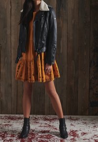 Czarna skórzana kurtka motocyklowa z owczą wełną przy kołnierzu, w połączeniu z sukienką w musztardowym kolorze z kwiatowym haftem i rozkloszowanym dołem. Czarne botki za kostkę.