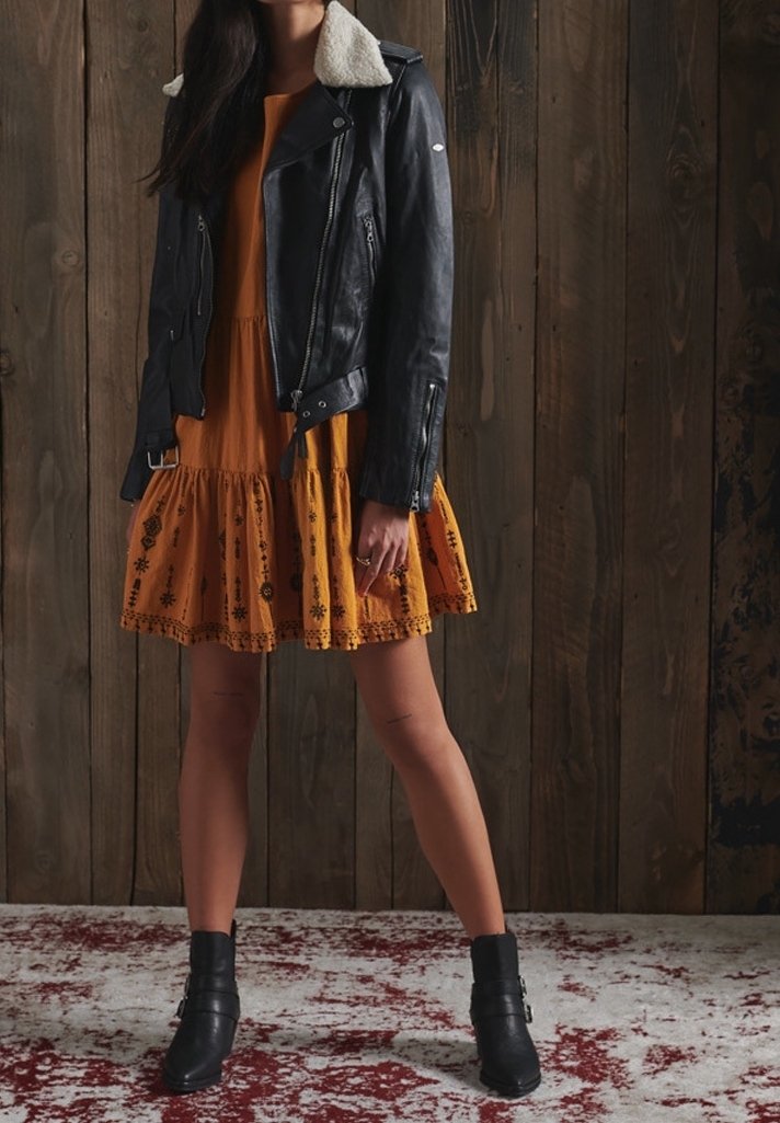 Czarna skórzana kurtka motocyklowa z owczą wełną przy kołnierzu, w połączeniu z sukienką w musztardowym kolorze z kwiatowym haftem i rozkloszowanym dołem. Czarne botki za kostkę.