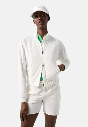 Kobieta w białej pikowanej kurtce zapinanej na zamek, dopasowanych spodenkach, białej czapce i zielonej koszulce pod spodem, stoi z jedną ręką w kieszeni.