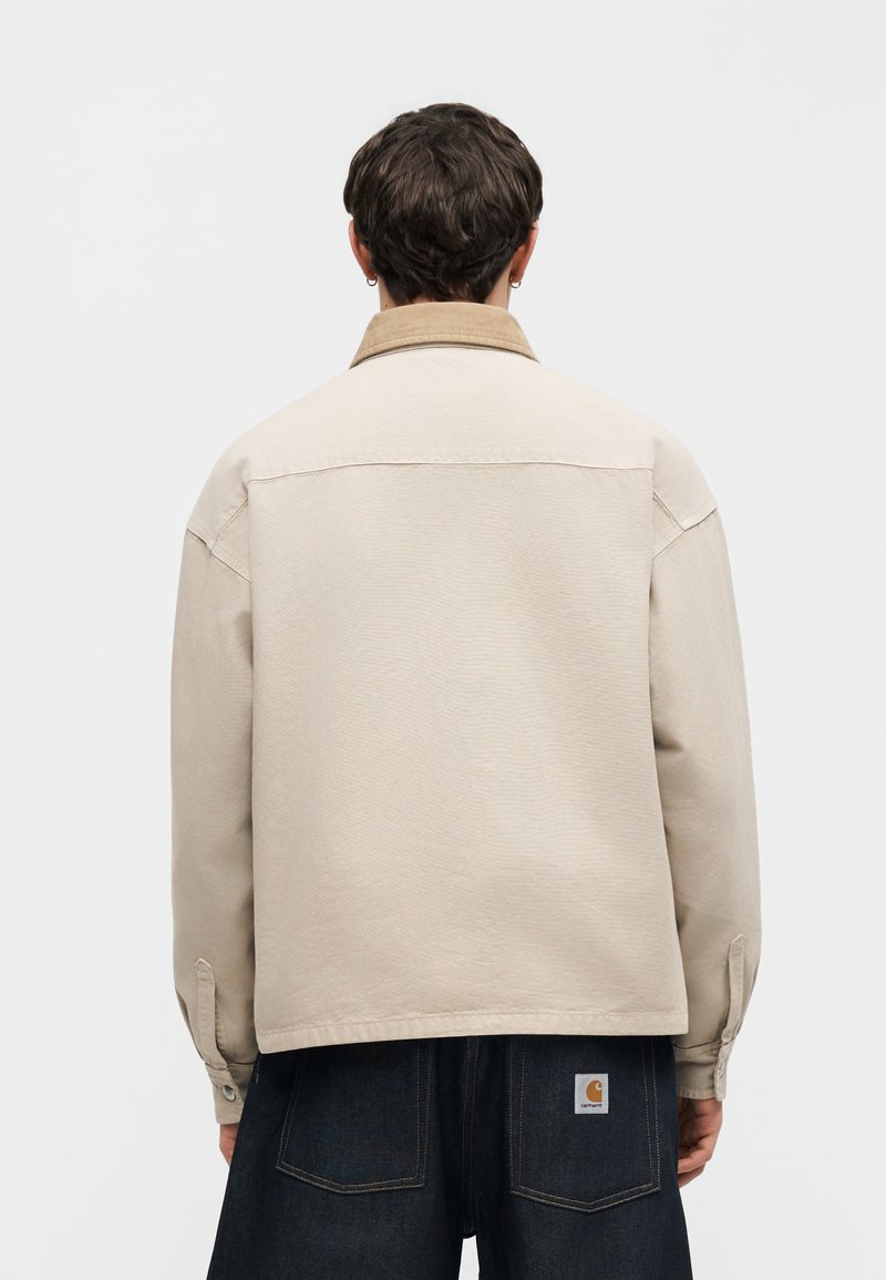 Joven con cabello corto y oscuro, vestido con una chaqueta beige con cuello marrón y vaqueros azul oscuro, de espaldas.