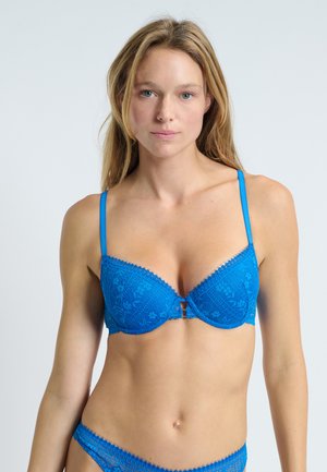Donna con lunghi capelli biondi che indossa un completo coordinato di reggiseno e slip in pizzo blu brillante, di fronte a uno sfondo chiaro e semplice.