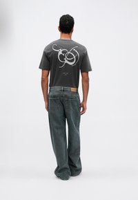 Camiseta gris con gráficos abstractos blancos en la parte posterior, combinada con unos vaqueros de denim sueltos de color verde oscuro. Ambas prendas tienen un corte holgado.