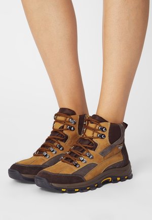 Botas de senderismo en marrón y beige con un diseño de caña alta, que cuentan con ojales de metal, cordones marrones, suelas de goma y detalles de malla.