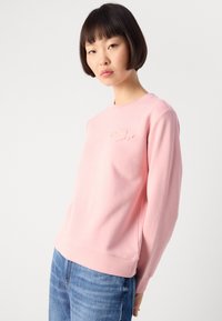 Jonge vrouw met kort donker haar, gekleed in een lichtroze sweatshirt met borduursel en blauwe jeans, staande tegen een plain witte achtergrond.