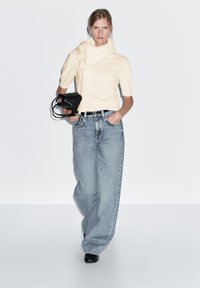 Maglione color crema con colletto arricciato, abbinato a jeans a gamba larga blu chiaro. Tiene in mano una piccola borsa nera, le scarpe nere completano il look.