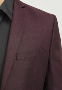 Bordeauxfarbener Blazer aus strukturiertem Stoff, mit einem Reverskragen und einer einzelnen Brusttasche, kombiniert mit einem schwarzen Hemd darunter.