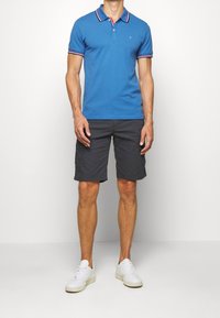 Uomo con polo a maniche corte blu, pantaloni cargo grigi scuri fino al ginocchio e sneaker bianche in piedi di fronte a uno sfondo bianco.