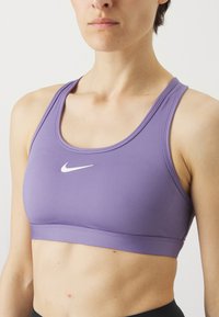 Ljubičasti sportski brus s mekim, rastezljivim materijalom i zaobljenim izrezom. Sadrži bijeli Nike logotip sprijeda, minimalističkog dizajna.