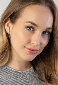 Boucle d'oreille en or avec une surface texturée, portée à l'oreille gauche. Gros plan sur une femme avec de longs cheveux châtain clair et un haut argenté.