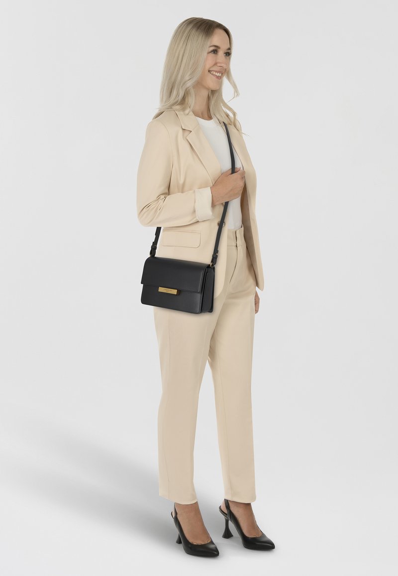 Black leather shoulder bag with a flap, gold hardware accent. Model wearing light beige suit, holding the bag with a long strap.
