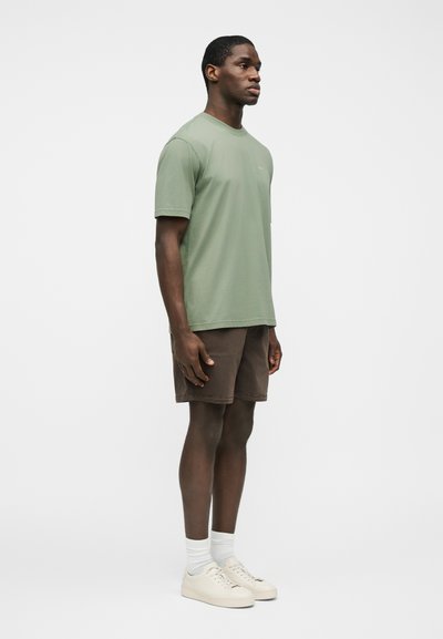 Άνδρας που στέκεται πλάγια, φοράει πράσινο πουκάμισο χρώματος φασκόμηλου, καφέ σορτς, λευκές κάλτσες και υπόλευκα αθλητικά παπούτσια, με φόντο απλό λευκό.