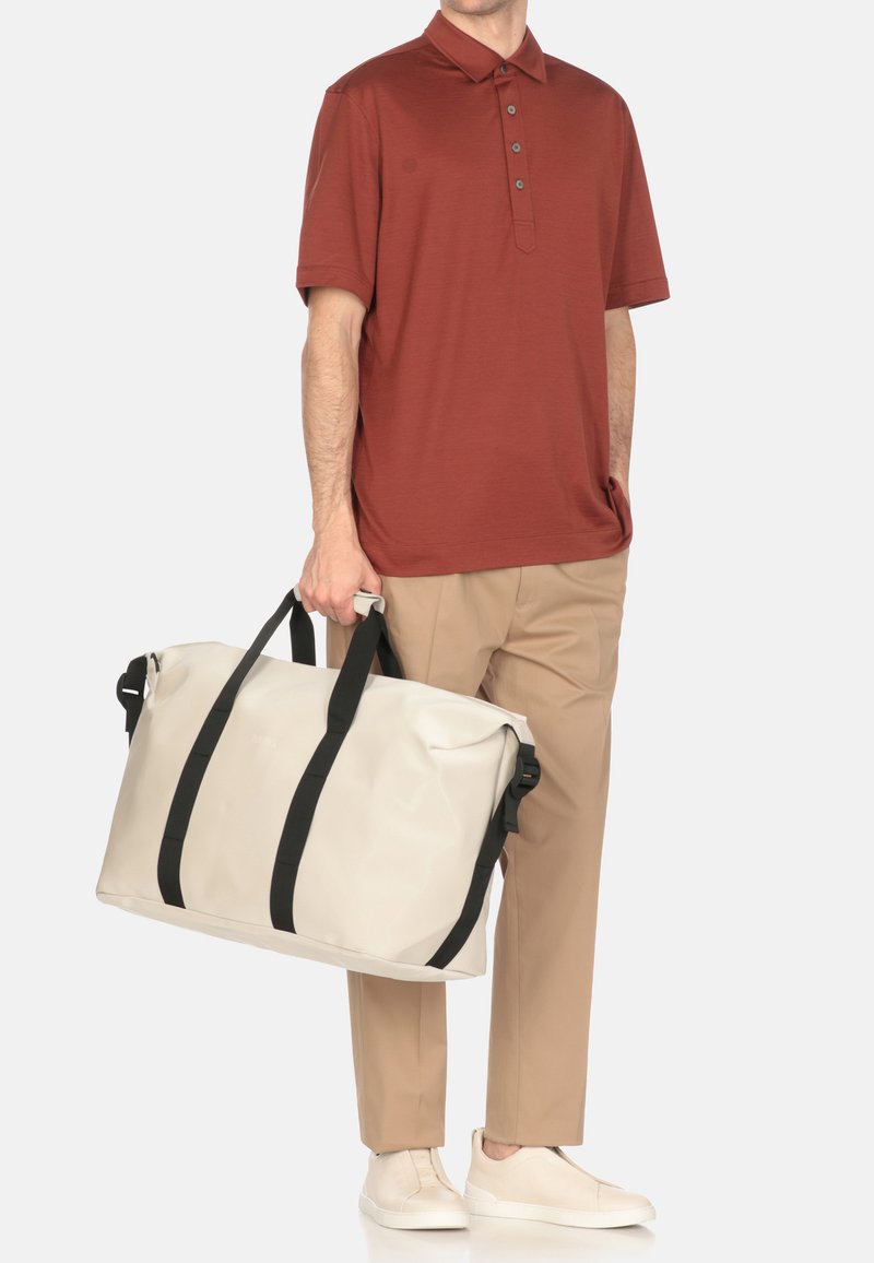 Beige waterproof duffel bag with black straps, held by a person wearing a rust-colored polo shirt and tan trousers, off-white slip-on shoes.