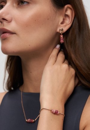 Frau trägt passende pinkfarbene Perlenohrringe und Armband mit goldenen Ketten, berührt ihren Hals, gekleidet in ein ärmelloses marineblaues Oberteil.