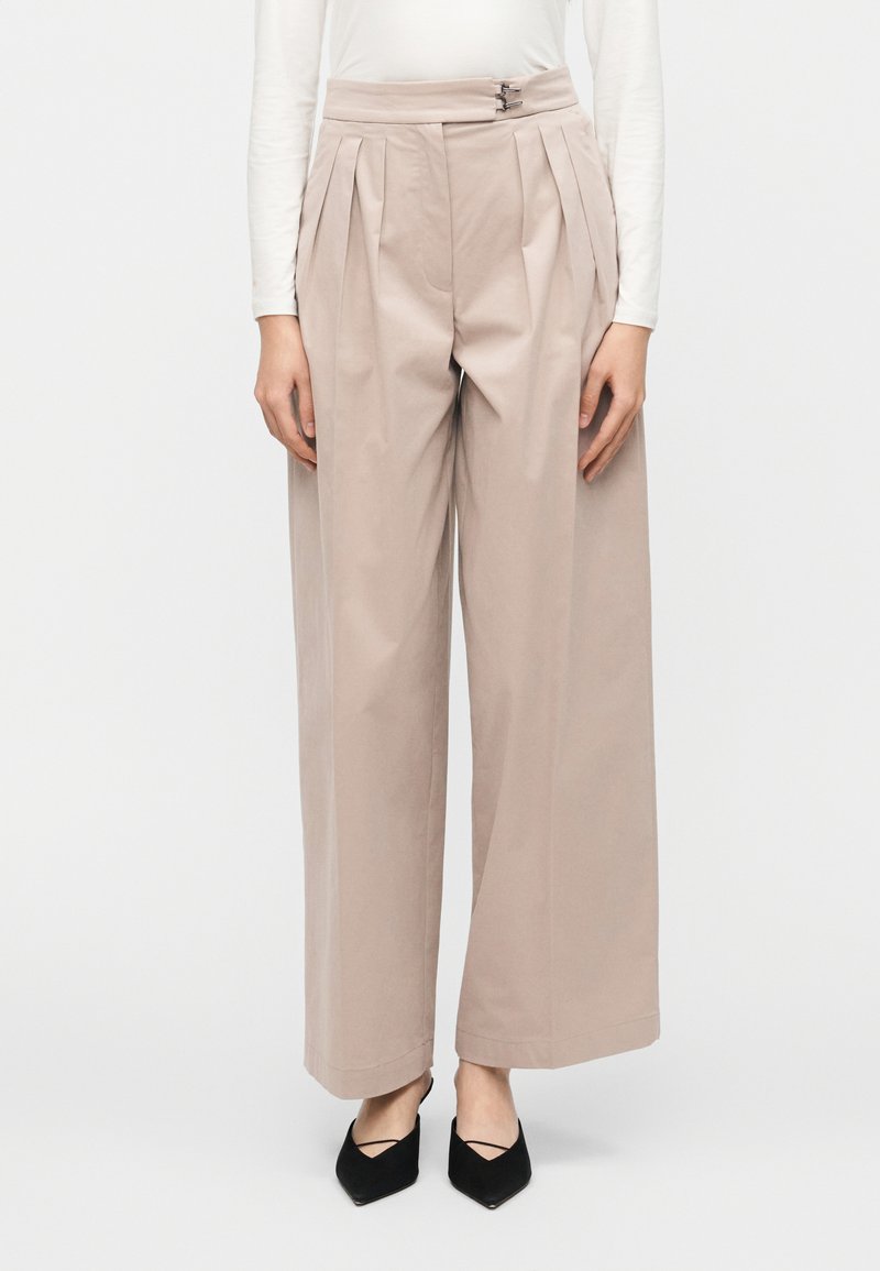 Μπεζ παντελόνι φαρδιάς γραμμής με πιέτες και ψηλ ή μέση. Διαθέτει μεταλλικά στοιχεία στη μέση. Λείο βαμβακερό ύφασμα, εφαρμοστή γραμμή.