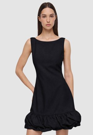 Donna con capelli scuri e lisci, che indossa un abito nero senza maniche con orlo arricciato a balze, in piedi davanti a uno sfondo chiaro e semplice.