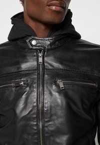 Schwarze Lederjacke mit einem Frontreißverschluss, zwei Brusttaschen mit Reißverschluss und einem schwarzen Hoodie-Futter. Glatte Textur mit Nähdetails.