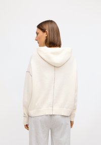 Cream-colored hoodie with a relaxed fit, textured fabric, hood, and a contrasting seam down the back. Paired with gray casual pants.