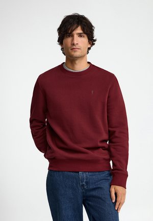 Hombre con cabello rizado y oscuro, vistiendo una sudadera granate y vaqueros azules, de pie frente a un fondo blanco liso.