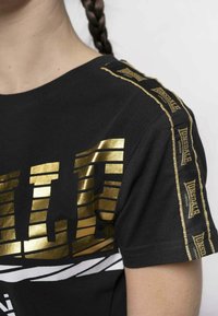 Black t-shirt featuring bold gold and white stripes across the chest, with gold logo accents along the shoulder seams. Soft cotton material.