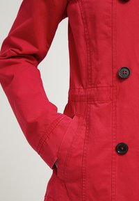 Chaqueta roja con textura suave, que presenta un cierre de botones, dos bolsillos laterales y detalles de costura prominentes.